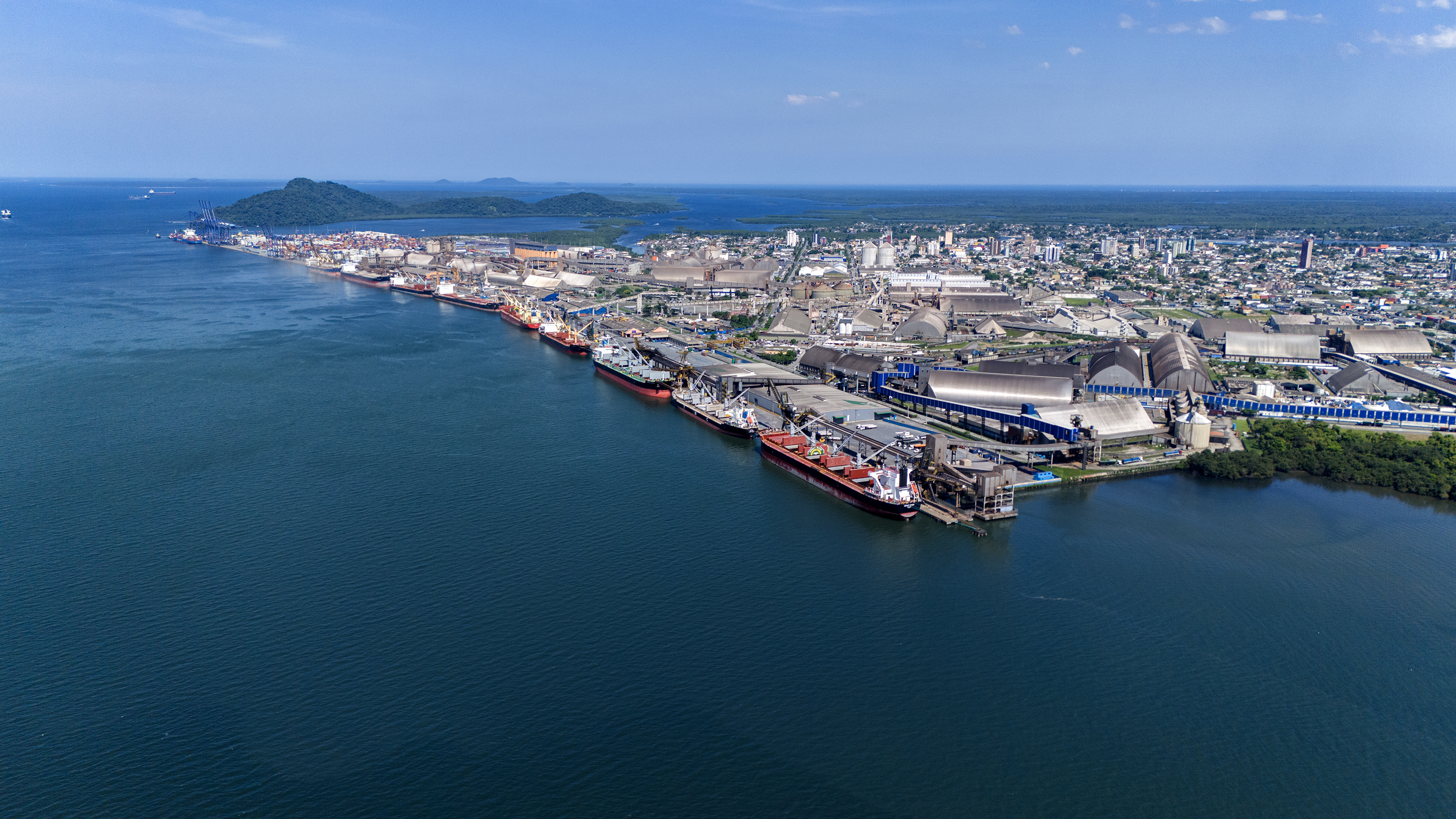 A imagem mostra o Porto de Paranaguá visto do alto, com uma grande faixa da Baía de Paranaguá na parte de baixo da foto até o final do porto. Os navios estão pequenos e enfileirados, a maioria na cor vermelha.