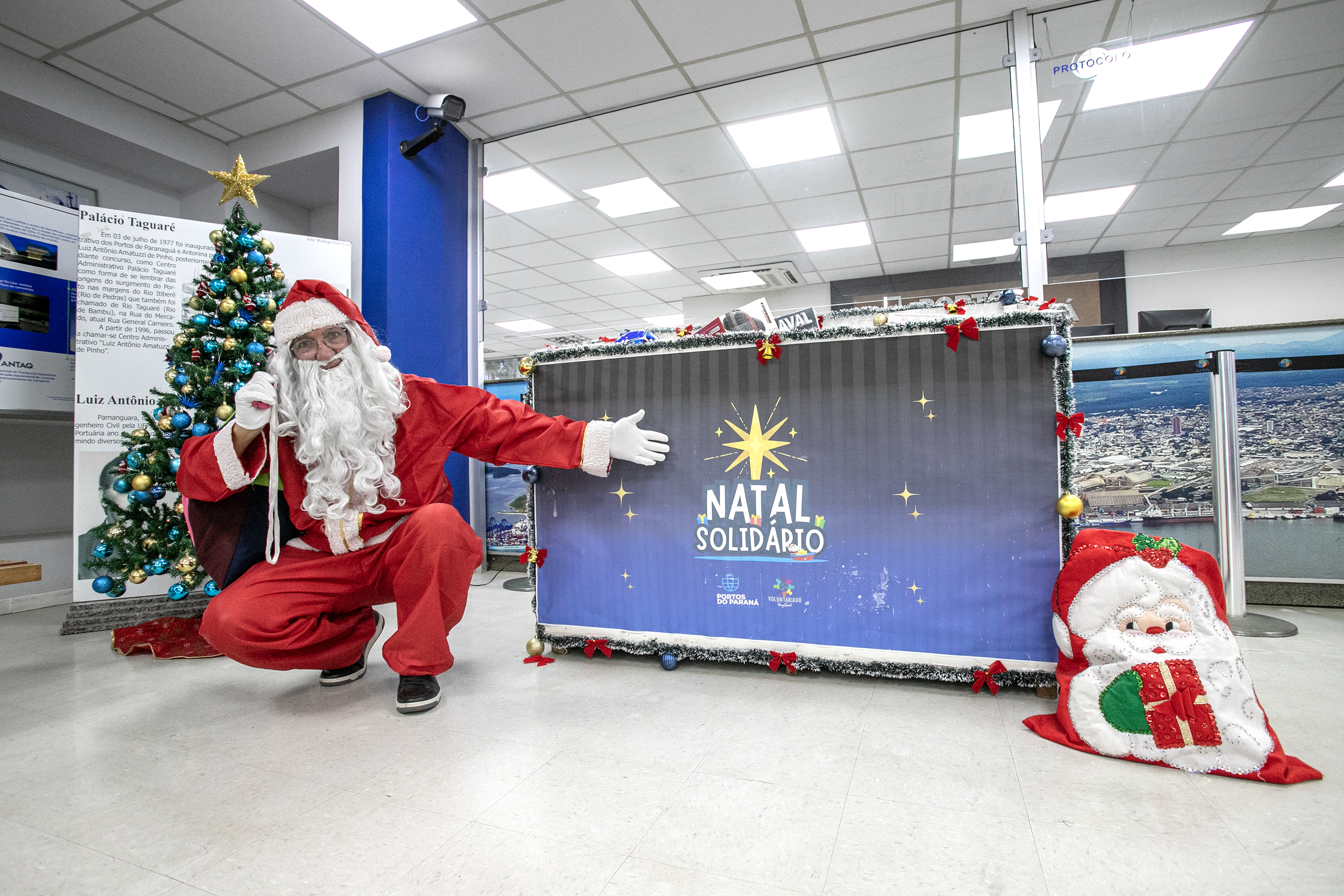 #ParaQuemNaoPodeVer A imagem mostra um homem vestido de Papai Noel agachado ao lado de uma árvore de Natal decorada com bolas azuis e douradas e uma estrela dourada no topo. Ele está usando um traje vermelho com detalhes brancos, incluindo um gorro e uma longa barba branca. O homem está fazendo um gesto de