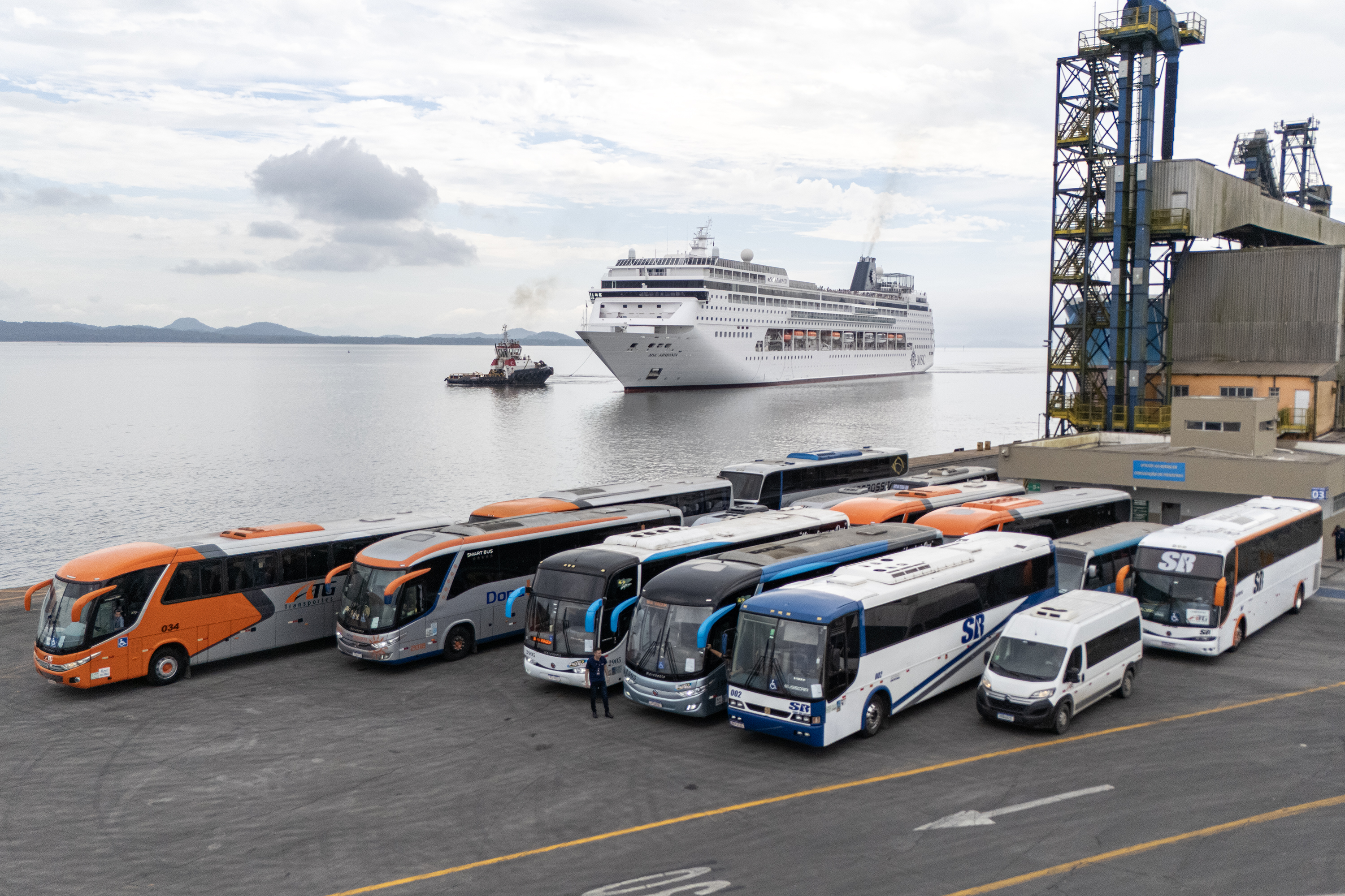 #praquemnaopodever 7 ônibus e uma van estão a frente da foto, no cais do Porto de Paranaguá, aguardando a chegada, atrás, do imponente navio de cruzeiros MSC Armonia.
