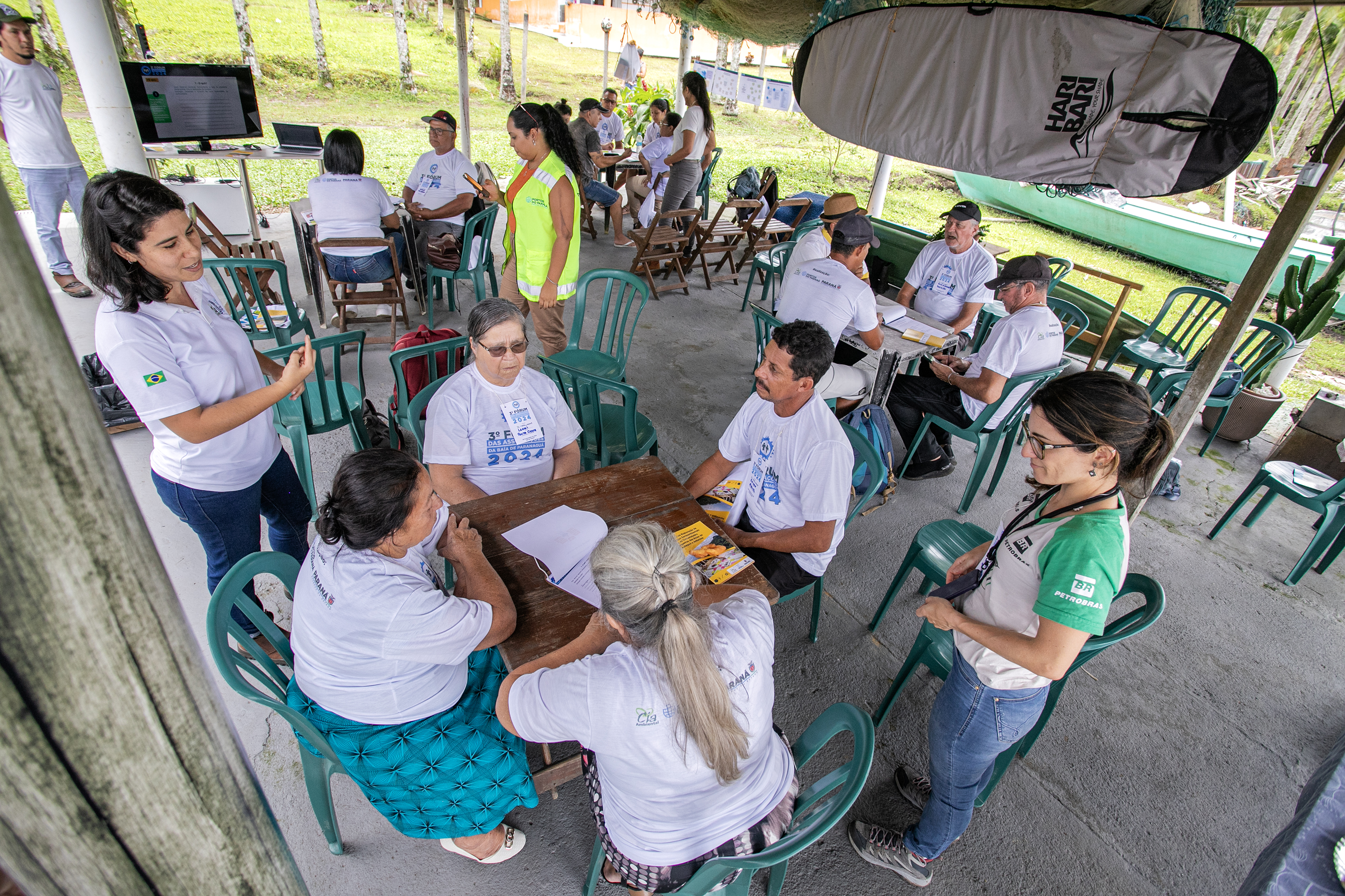 #praquemnaopodever várias pessoas sentadas em cadeiras de plástico verdes separadas em grupos gesticulam sobre projetos socioambientais. Todos vestem camisas brancas e conversam sérios enquanto outras pessoas em pe passam pelas pesas. Um prancha está presa ao teto e ao fundo é possível vera grama da Ilha do Teixeira