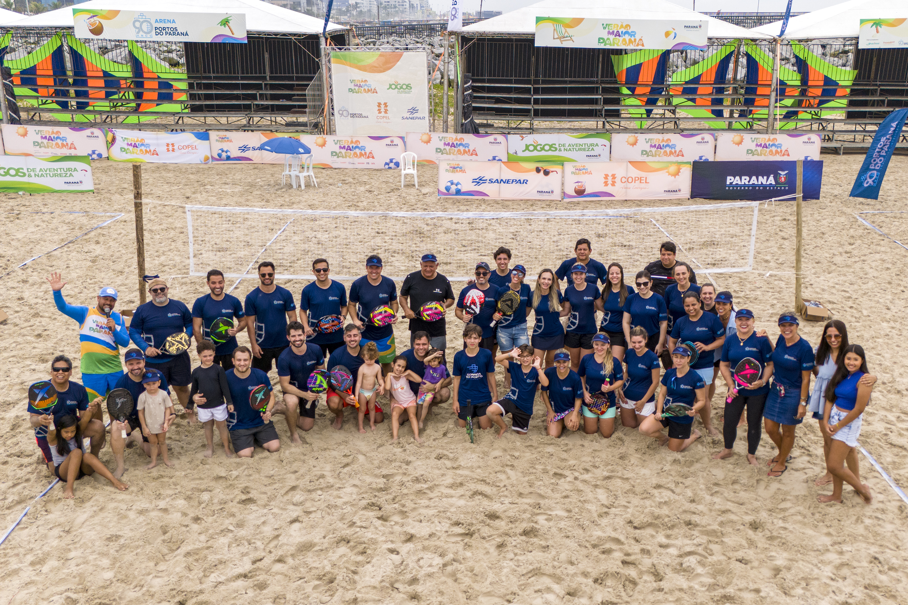 #ParaQuemNaoPodeVer A imagem mostra um grupo grande de pessoas posando para uma foto em uma quadra de areia, possivelmente de vôlei de praia. Todos estão vestindo camisetas azuis e alguns seguram bolas coloridas. Ao fundo, há uma estrutura com várias faixas e banners, que parecem ser de patrocinadores ou eventos, com logotipos e textos.