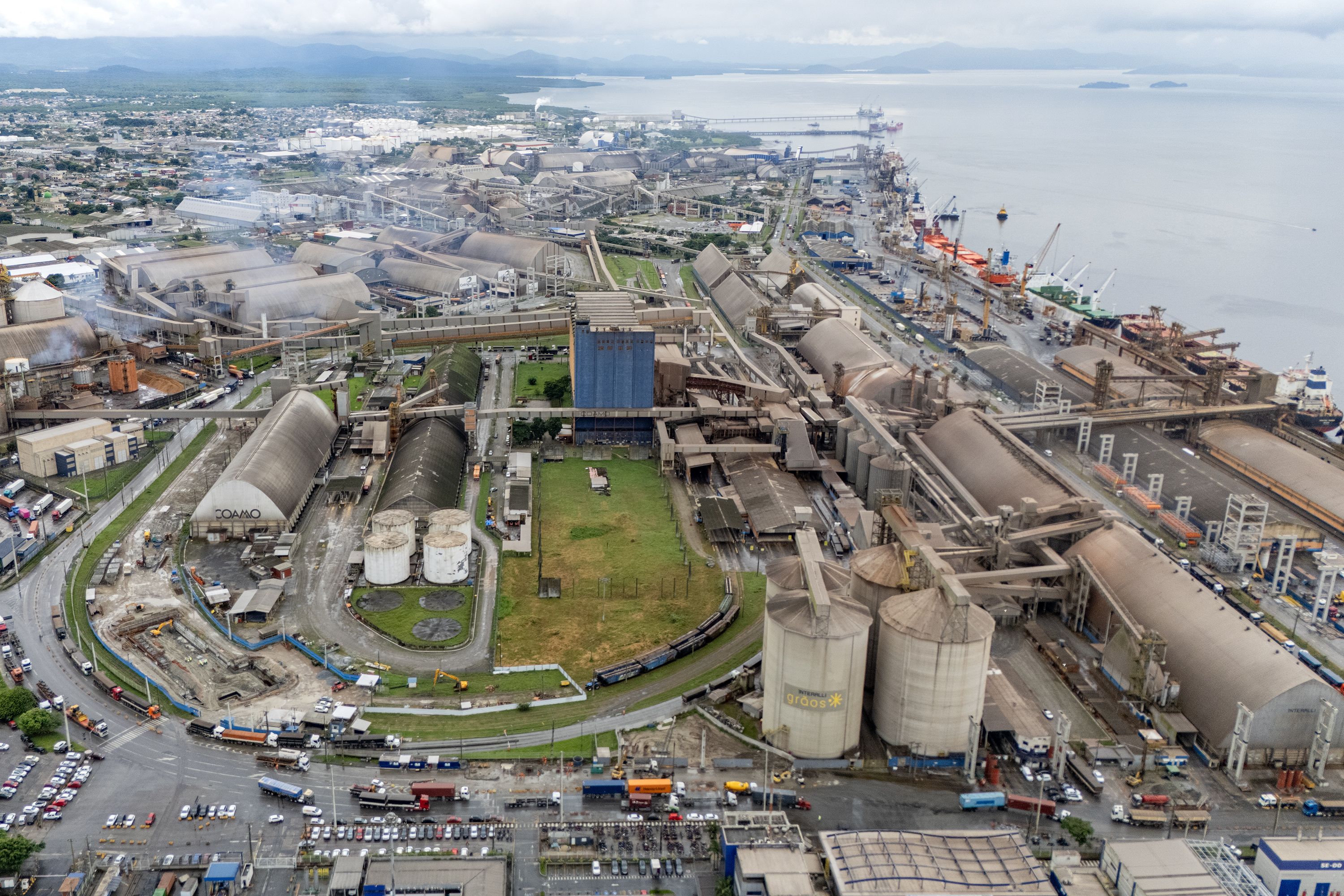 #ParaQuemNaoPodeVer A imagem mostra uma foto aérea de uma área portuária. Na imagem, é possível ver várias estruturas grandes, como tanques cilíndricos e prédios industriais, além de áreas verdes ao redor. Há também várias estradas e veículos visíveis.