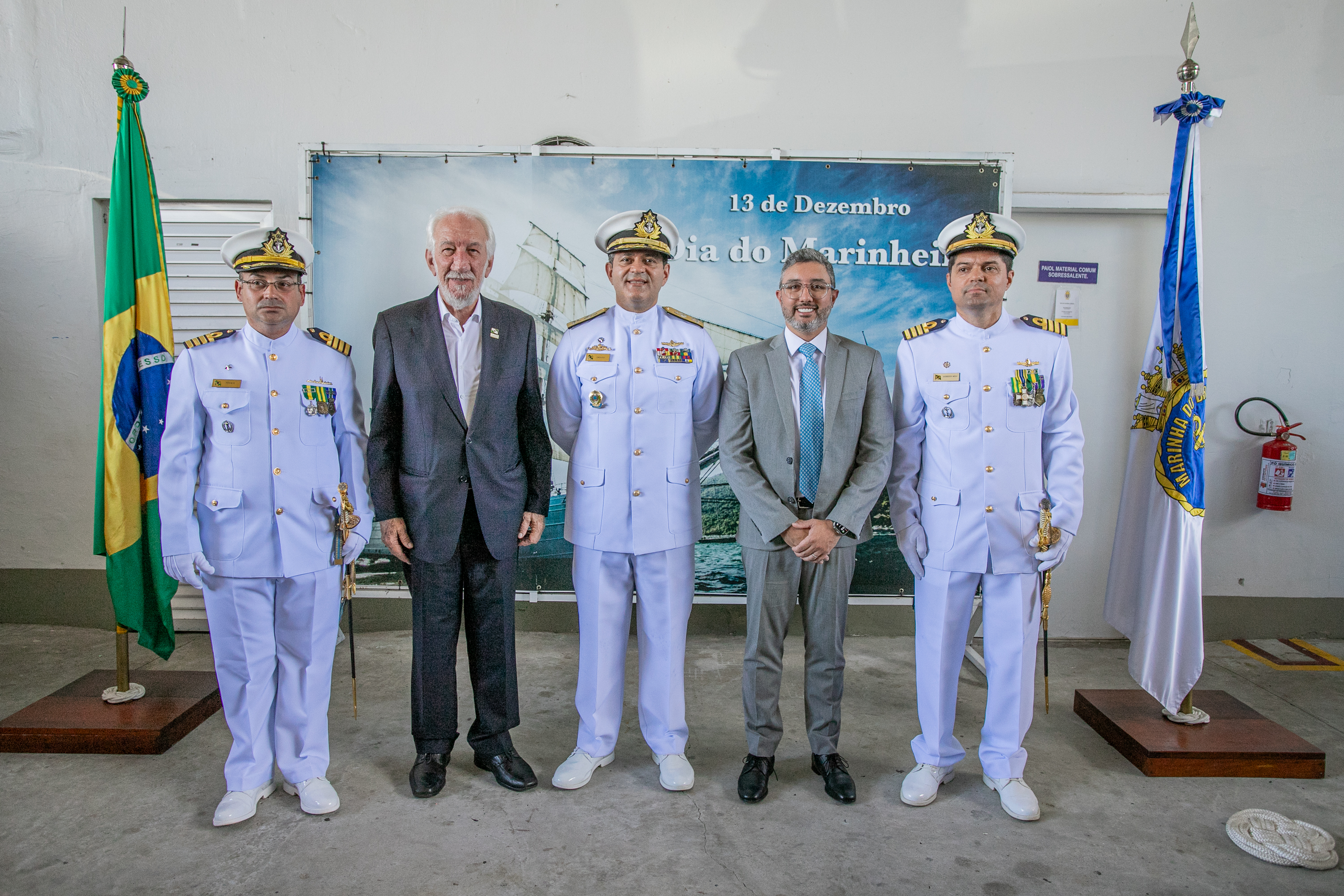 #ParaQuemNaoPodeVer A imagem mostra cinco homens posando para uma foto em frente a uma bandeira do Brasil e uma bandeira da Marinha do Brasil. Três deles estão vestidos com uniformes brancos de oficiais da Marinha, com chapéus brancos e insígnias no peito. Os outros dois homens estão usando ternos. Ao fundo, há um banner azul com o texto
