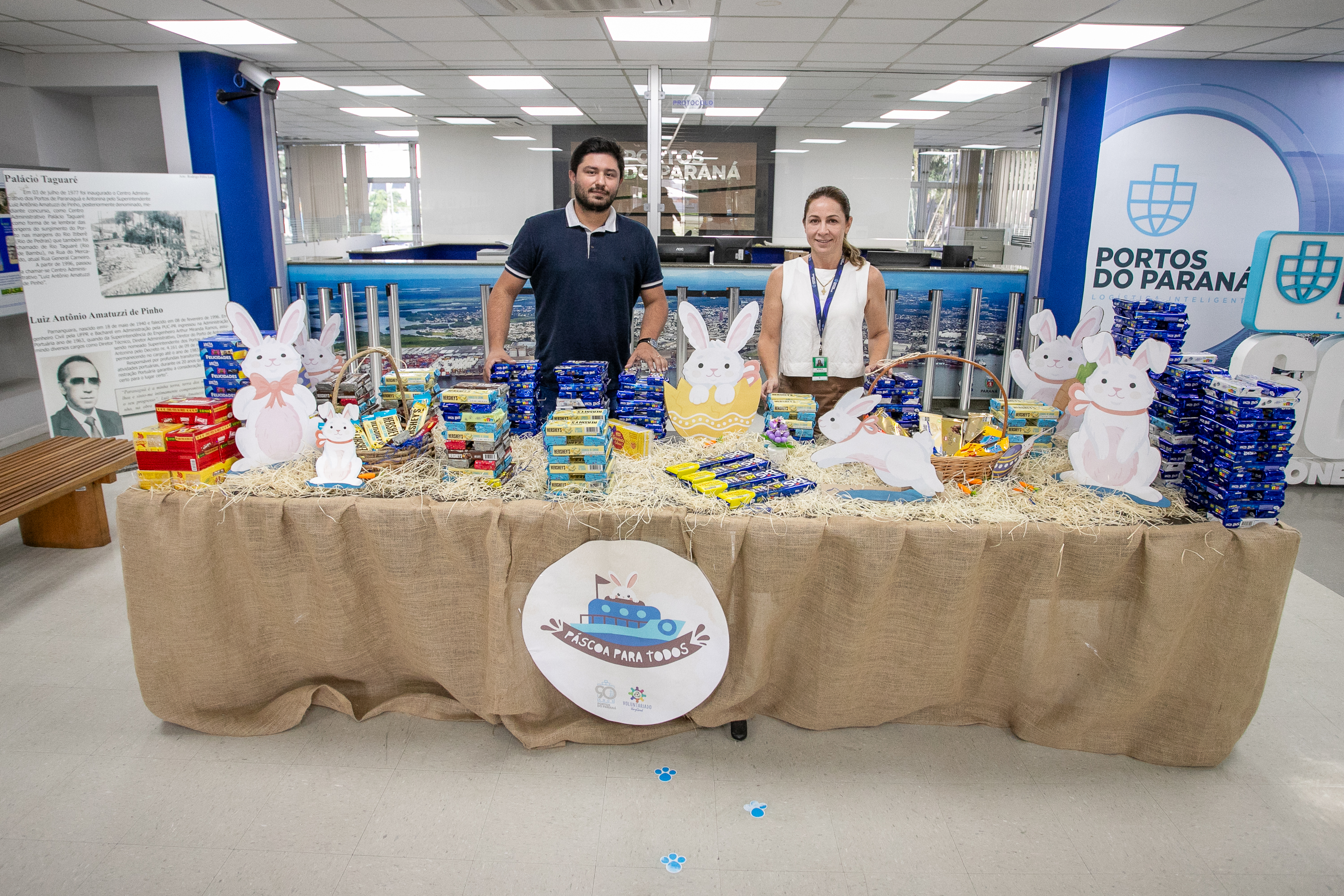 #ParaQuemNaoPodeVer A imagem mostra uma mesa coberta com uma toalha de tecido bege, decorada com coelhos de Páscoa e diversos produtos organizados em cima, como caixas de chocolate. Duas pessoas estão atrás da mesa, um homem e uma mulher, ambos sorrindo. Ao fundo, há um banner com o logotipo