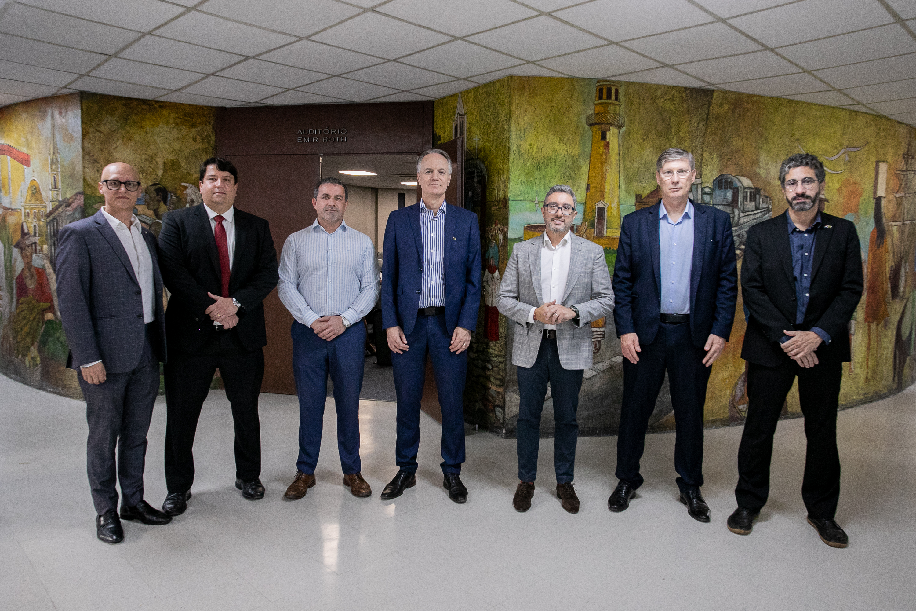 #ParaQuemNaoPodeVer Imagem exibe uma imagem de seis homens em pé, lado a lado, em um ambiente interno. Eles estão vestidos formalmente, com ternos e camisas sociais. Ao fundo, há uma parede com uma pintura colorida.