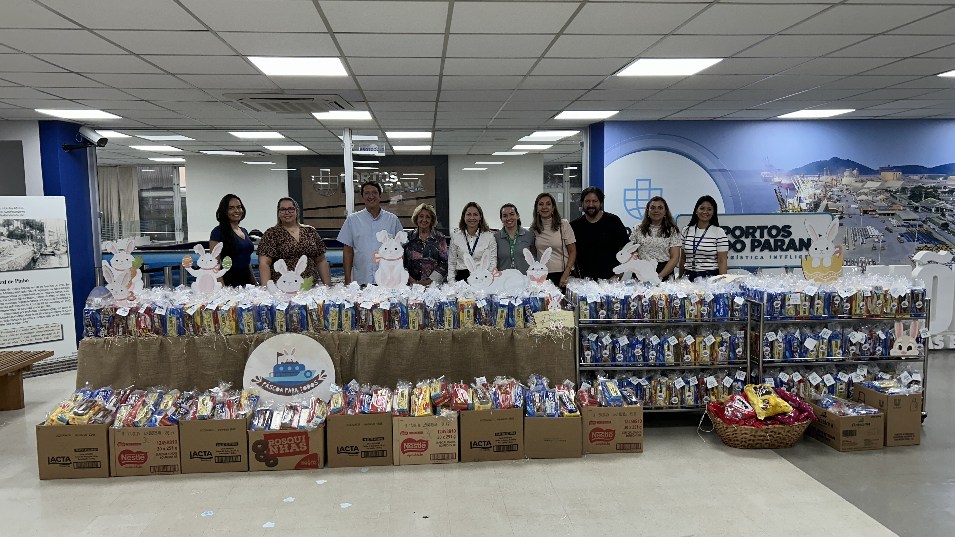 #ParaQuemNaoPodeVer A imagem exibe um grupo de pessoas em pé atrás de uma mesa cheia de cestas ou pacotes embrulhados, possivelmente para doação. As cestas estão decoradas com coelhos de papel, sugerindo uma temática de Páscoa. Há várias caixas de produtos empilhadas ao lado das cestas. Na parede ao fundo, há um logotipo azul e branco com um desenho que parece ser um edifício ou estrutura. As pessoas estão sorrindo e posando para a foto.