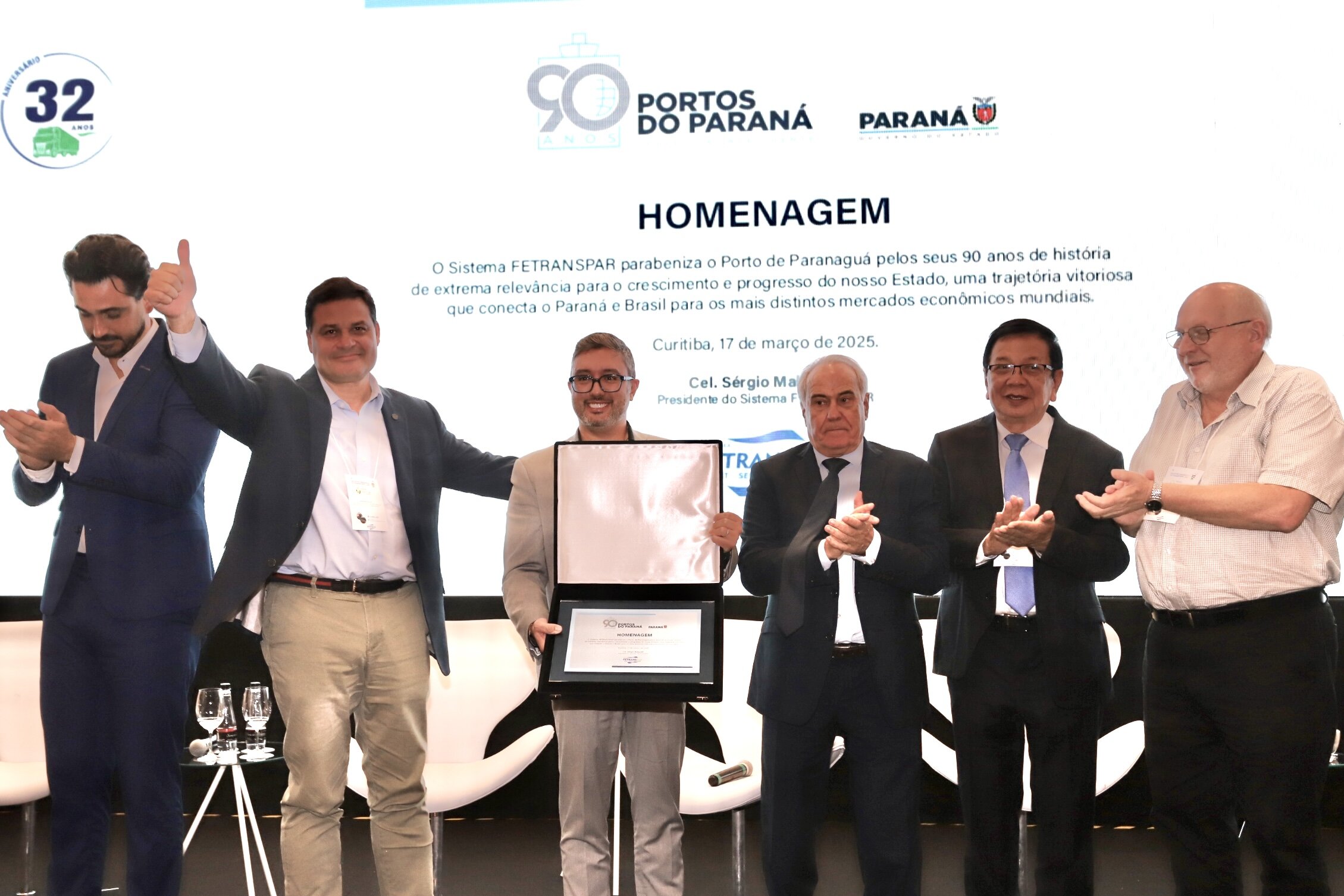 #praquemnaopodever homem te terno cinza e óculos de grau segura uma caixa aberta com uma placa dentro. Ele e outros cinco homens brancos de roupa social estão em cima de um palco, alguns sorriem, outroas fazem joinha. Ao fundo, aparece escrito