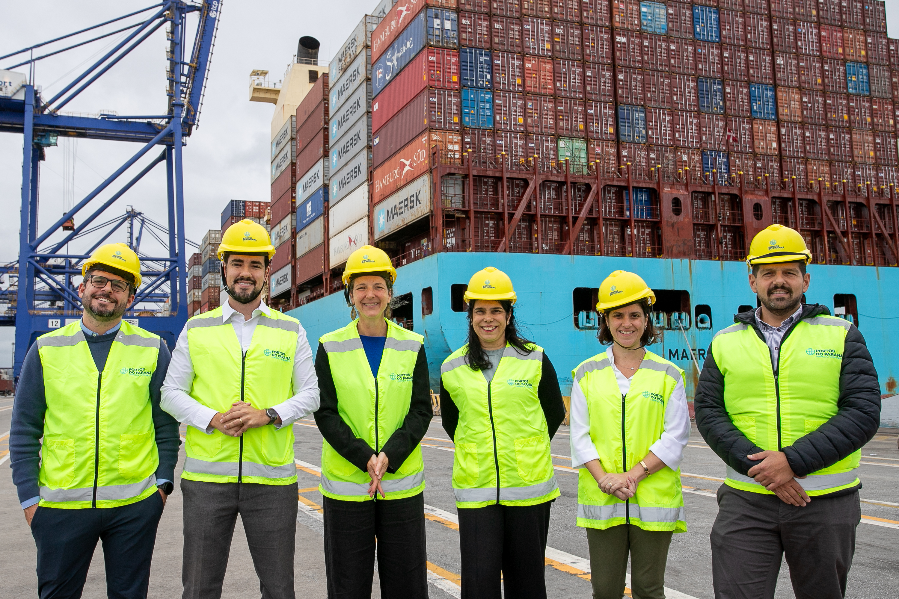 #ParaQuemNaoPodeVer Seis pessoas estão posando para uma foto em frente a um cenário de contêineres empilhados em um porto. Todos estão usando coletes de segurança amarelos e capacetes amarelos. Ao fundo, há uma estrutura de guindaste azul e os contêineres têm várias cores e logotipos, incluindo