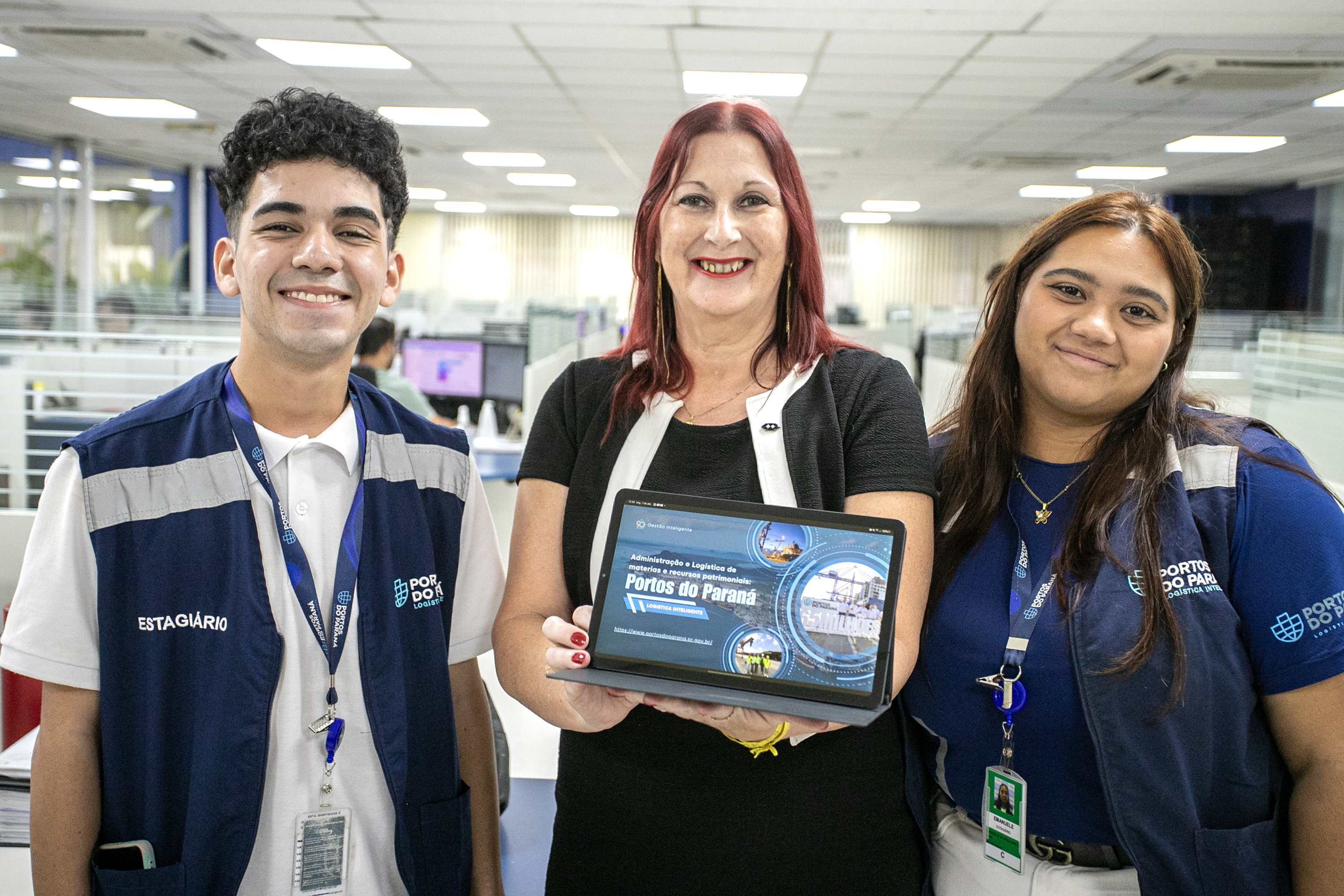 #ParaQuemNaoPodeVer Três pessoas estão sorrindo para a câmera em um ambiente interno, possivelmente um escritório. A pessoa à esquerda é um jovem de cabelo curto e escuro, vestindo um colete azul com a palavra