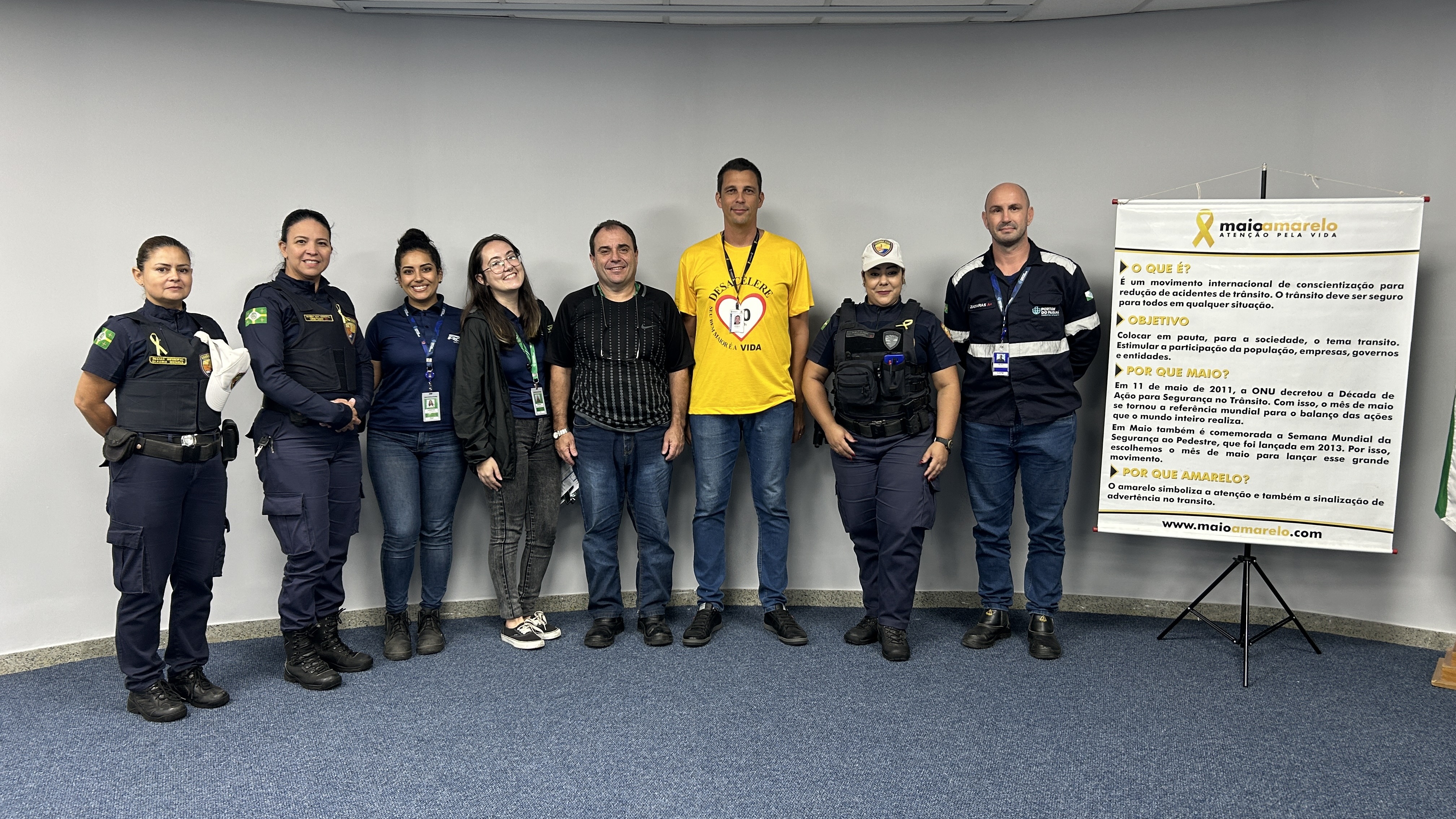 #ParaQuemNaoPodeVer Foto de um grupo de oito pessoas, sendo quatro delas usando uniformes de agentes de trânsito ou policiais, e as outras quatro usando roupas casuais. Eles estão em pé, lado a lado, em frente a uma parede clara e um tapete azul. À frente do grupo, há um banner com informações sobre o movimento Maio Amarelo, que é uma campanha de conscientização para a segurança no trânsito. O banner tem detalhes em amarelo e branco, com o símbolo do laço amarelo. Todos estão sorrindo e olhando para a câmer