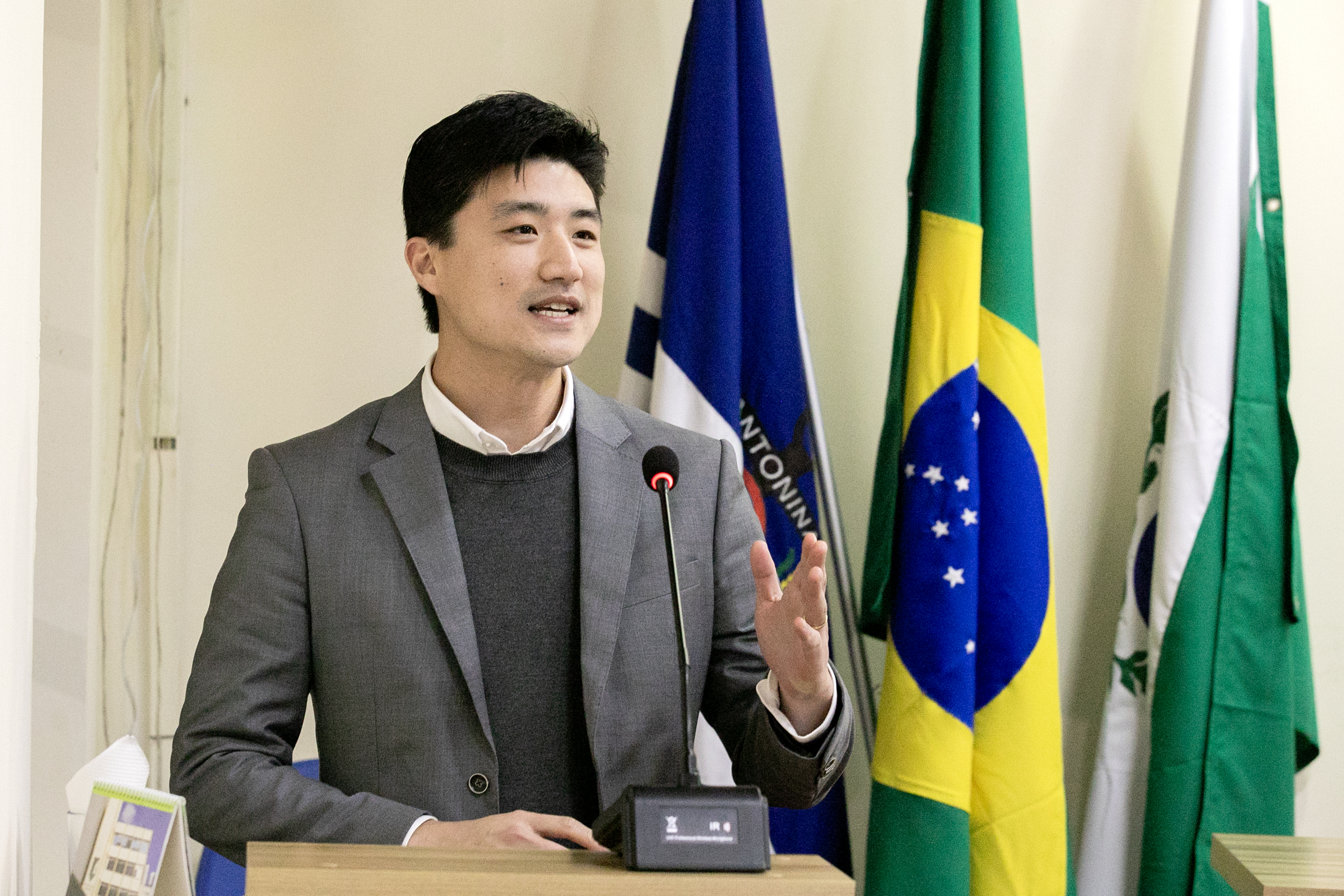 #ParaQuemNaoPodeVer foto de um homem jovem, de pele clara e cabelo preto, vestindo terno cinza, suéter escuro e camisa branca. Ele está em pé atrás de um púlpito, falando ao microfone, com expressão sorridente. Atrás dele, há bandeiras, incluindo a do Brasil e outra azul com a palavra