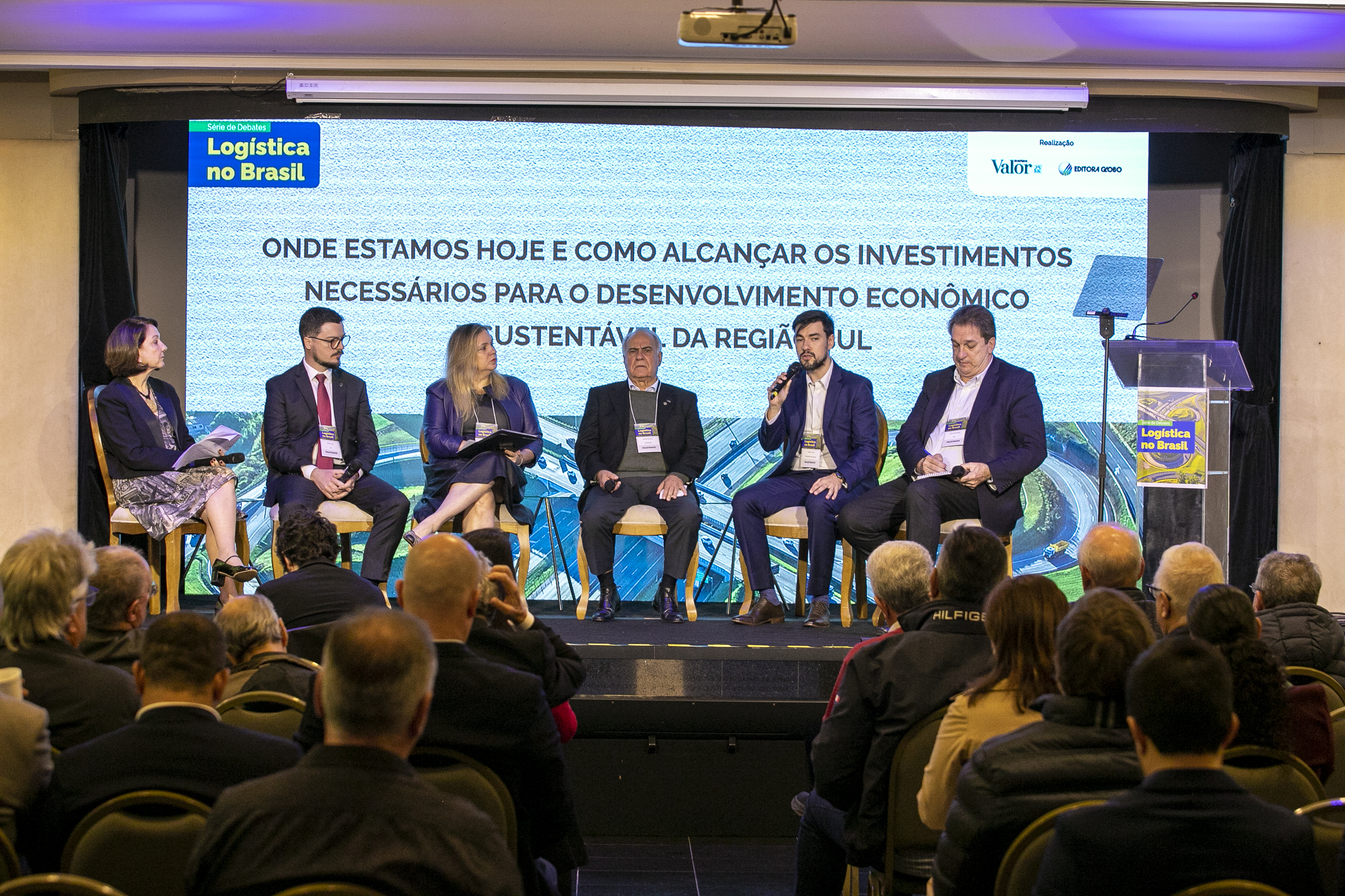 #ParaQuemNaoPodeVer Foto de um painel em um evento corporativo, com cinco pessoas (quatro homens e uma mulher) sentadas no palco, participando de um debate. No fundo, há um grande telão com o texto:
