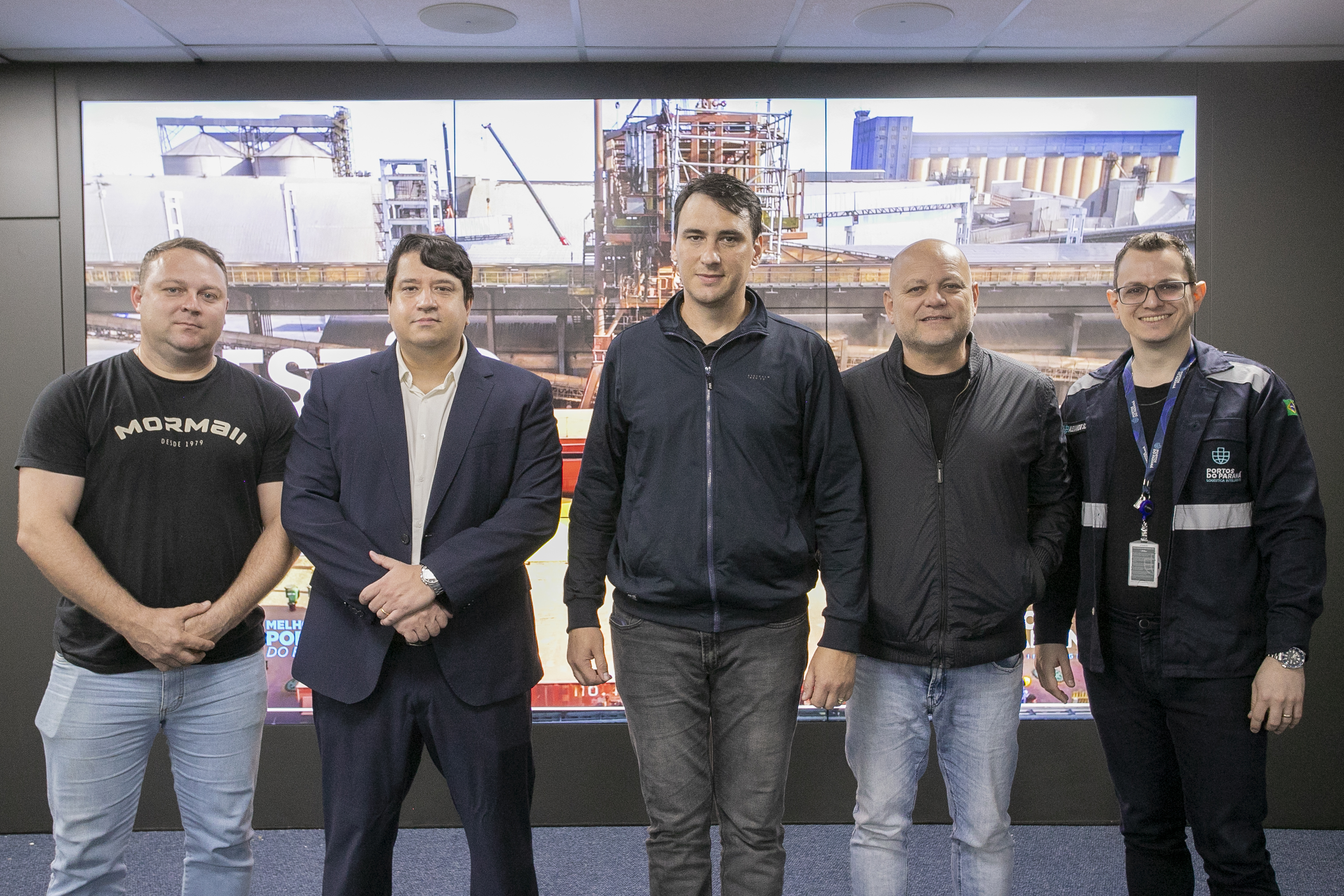 #ParaQuemNaoPode Ver Imagem mostra cinco homens posando para uma foto em grupo. Eles estão em pé, lado a lado, com um fundo que parece ser uma imagem de uma área industrial ou de construção, com estruturas metálicas e guindastes. Um dos homens, na parte superior esquerda, veste uma camiseta preta com a palavra