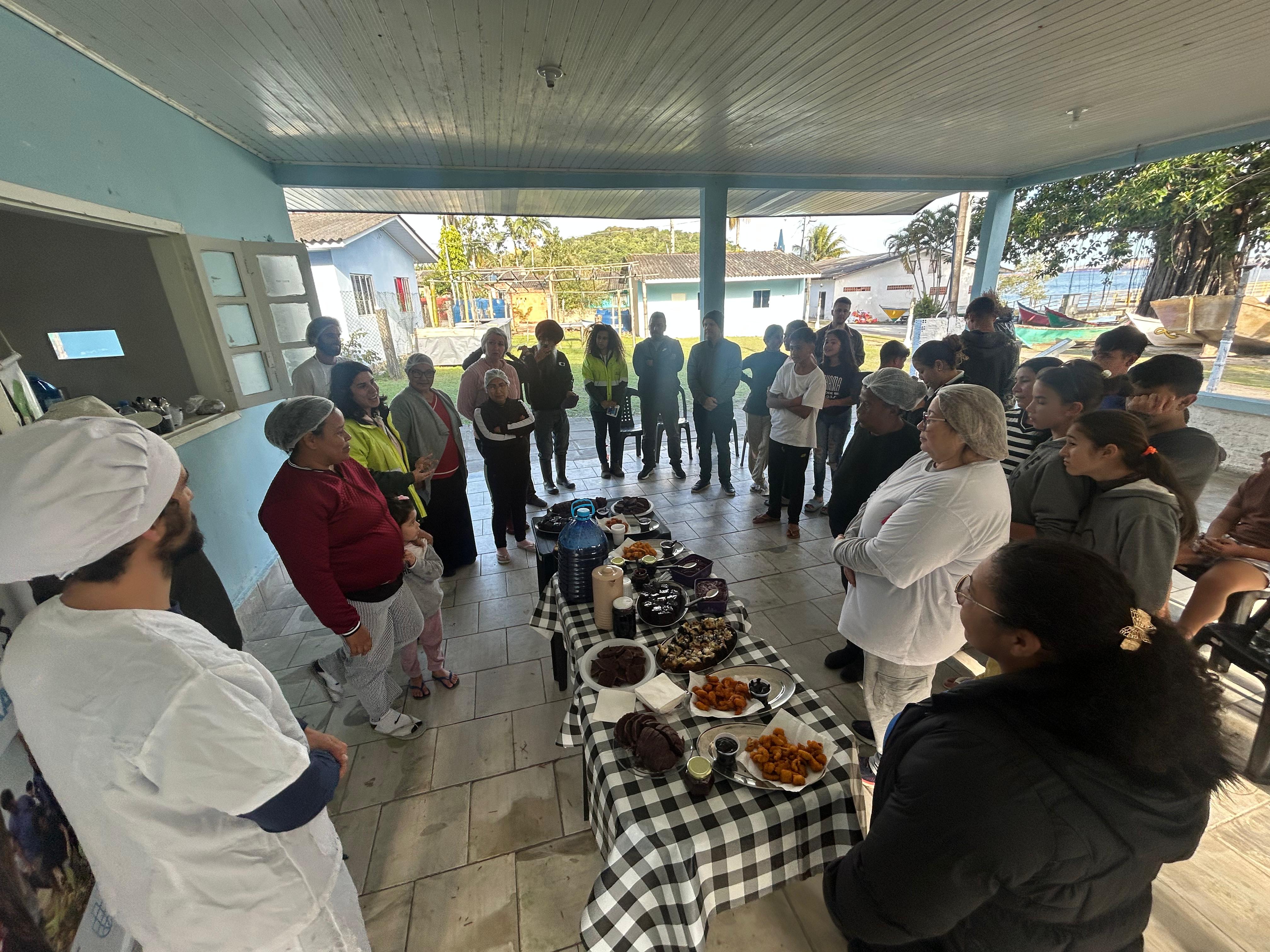 #praquemnaopodever foto mostra um grupo de pessoas em círculo em volta de uma mesa com toalha xadrez e repleta de comida. A maioria olha para uma pessoa que fala no outro lado da foto.