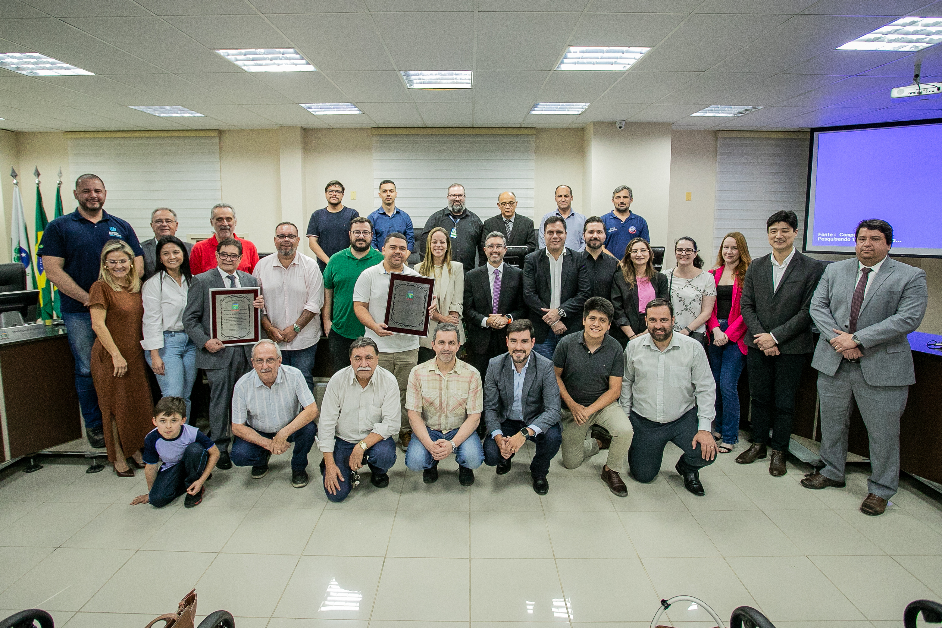 #ParaQuemNaoPodeVer Foto de um grupo grande de pessoas em um ambiente fechado, possivelmente uma sala de reuniões ou auditório. Todos estão vestidos de maneira formal e informal, sugerindo um evento corporativo ou de premiação. Algumas pessoas seguram placas ou certificados, indicando que receberam algum tipo de reconhecimento. Ao fundo, há bandeiras, reforçando o clima institucional do local. As pessoas estão sorrindo e posando para a foto, aparentando clima de celebração ou comemoração.