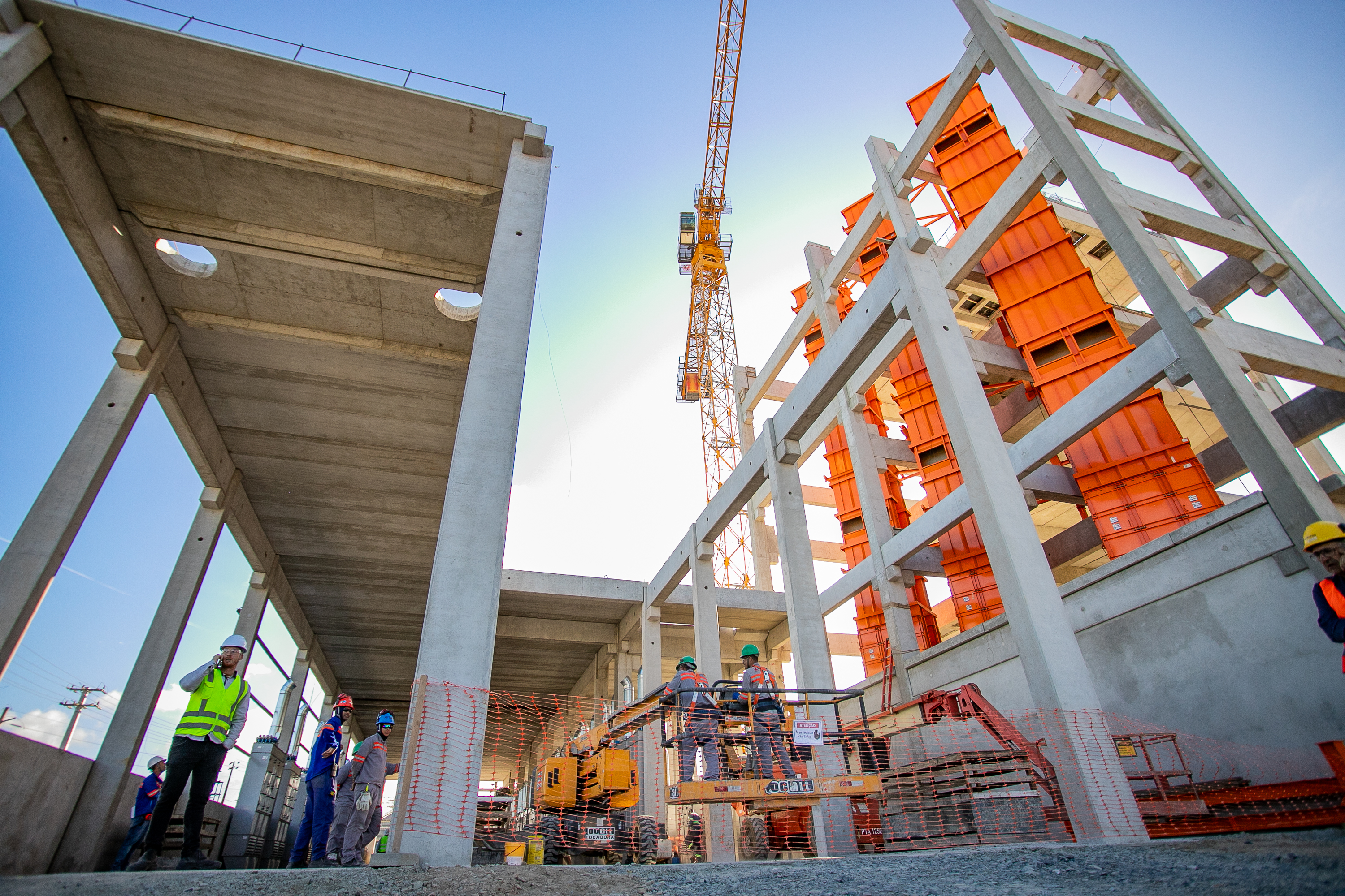 #praquemnaopodever foto do chão do canteiro de obras do Moegão, com os pilares cinzas e máquinas laranjas, trabalhadores estão no local espalhados trabalhando e o céu está de fundo, em um azul claro.