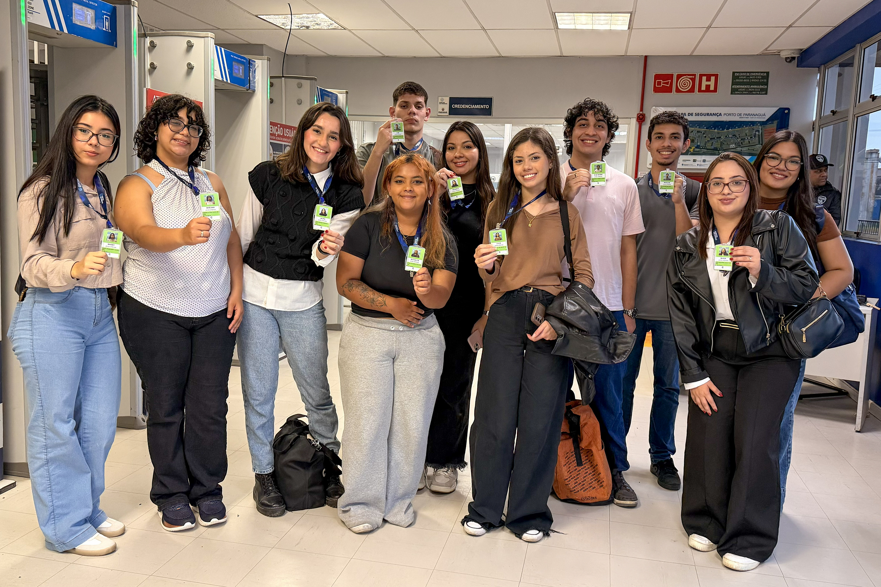 #ParaQuemNaoPodeVer Grupo de onze jovens alinhados em um corredor interno, todos segurando crachás verdes voltados para a frente. Na foto, as pessoas estão em pé, lado a lado, sorrindo. Há diversidade de tons de pele, de claros a médios e mais escuros, e estilos de cabelo diferentes, incluindo cabelos lisos, cacheados e ondulados, alguns longos, outros curtos, em cores como castanho, preto e loiro. As roupas são casuais: calças jeans claras e escuras, calça de moletom cinza, calça preta, tênis brancos