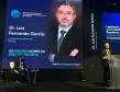 #ParaQuemNaoPodever A imagem mostra uma apresentação ou conferência. No centro da tela, há um slide com fundo azul escuro e texto em branco e azul claro. O texto diz "Dr. Luiz Fernando Garcia" e "Presidente da Portos do Paraná". Abaixo, está escrito "OS MAIORES NOMES DA ÁREA TRABALHISTA". No canto inferior direito do slide, há um logotipo circular azul com linhas horizontais.  Na parte inferior da tela, há uma foto de um homem de terno e gravata, que parece ser o Dr. Luiz Fe
