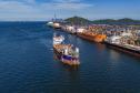 #ParaQuemNaoPodeVer A imagem mostra uma fotografia de um porto com vários barcos atracados. A água do mar é de um azul intenso e há uma grande quantidade de barcos coloridos, principalmente em tons de branco e azul, ao longo de um cais. Ao fundo, é possível ver uma linha de montanhas sob um céu azul claro.