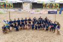 #ParaQuemNaoPodeVer A imagem mostra um grupo grande de pessoas posando para uma foto em uma quadra de areia, possivelmente de vôlei de praia. Todos estão vestindo camisetas azuis e alguns seguram bolas coloridas. Ao fundo, há uma estrutura com várias faixas e banners, que parecem ser de patrocinadores ou eventos, com logotipos e textos.