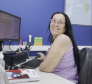 #praquemnaopodever Uma mulher de cabelos negros e lisos, regata lilas e óculos sorri para a camera. Ela está sentada em frente ao computador, mas precisou virar de lado para posar pra foto. O ambiente é de escritório.