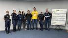 #ParaQuemNaoPodeVer Foto de um grupo de oito pessoas, sendo quatro delas usando uniformes de agentes de trânsito ou policiais, e as outras quatro usando roupas casuais. Eles estão em pé, lado a lado, em frente a uma parede clara e um tapete azul. À frente do grupo, há um banner com informações sobre o movimento Maio Amarelo, que é uma campanha de conscientização para a segurança no trânsito. O banner tem detalhes em amarelo e branco, com o símbolo do laço amarelo. Todos estão sorrindo e olhando para a câmer