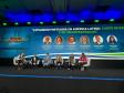 #ParaQuemNaoPodeVer Palco de conferência com seis pessoas sentadas em cadeiras, participando de um painel de discussão. Ao fundo, há um grande painel azul com a imagem de um contêiner portuário e o título do evento em espanhol: "Expansión portuaria en América Latina: casos de éxito y retos estratégicos". Abaixo do título, aparecem fotos e nomes dos participantes do painel, com seus respectivos cargos e empresas. O ambiente é formal, com todos vestidos de maneira profissional. O palco está bem iluminado e há