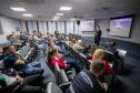 #ParaQuemNaoPodeVer Auditório com várias pessoas sentadas em cadeiras, todas de cabeça baixa. Na frente, um homem de terno está em pé, também de cabeça baixa, próximo a um púlpito transparente. Ao fundo, há dois telões projetando a mesma imagem, com fundo roxo e texto branco, mas o texto não está totalmente legível na foto. O ambiente é fechado, com iluminação de teto e paredes claras.