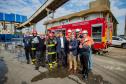#praquemnaopodever Na foto um grupo de homens faz o sinal de jóia com o polegar e sorriem em frente a um caminhão de bombeiros. Ao fundo, um predio branco e dalas suspensas que conectam empresas ao cais.
