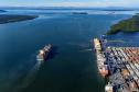 #ParaQuemNaoPodeVer Imagem aérea de um porto mostrando um grande navio saindo do cais, cercado de contêineres e equipamentos portuários. O mar, de azul intenso, ocupa a maior parte da imagem, com algumas pequenas ilhas verdes ao fundo. Outros navios menores podem ser vistos mais afastados, navegando ao longe.