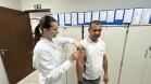 #ParaQuemNaoPodeVer Cena dentro de um consultório ou ambiente de saúde. Uma mulher de cabelo preso, óculos e jaleco branco está aplicando uma injeção no braço de um homem. O homem veste uma camiseta branca com a palavra "ENFERMAGEM" estampada. Eles estão em pé, próximos a armários brancos, com vários papéis presos em pastas azuis na parede ao fundo. Há também uma placa de saída de emergência verde na parede superior direita.