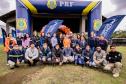 #ParaQuemNaoPodeVer Foto de um grupo grande de pessoas posando para a câmera, todas sorrindo, em frente a banners e bandeiras com os logotipos da Polícia Rodoviária Federal (PRF) e “Portos do Paraná – Logística. Eficiência. Valor.” Ao fundo, há balões laranja reunidos decorando a entrada de um prédio. As pessoas estão ao ar livre sobre uma área gramada, usam roupas variadas, incluindo uniformes da PRF, coletes e agasalhos. O ambiente é de evento institucional ou comemorativo, possivelmente uma ação conjunta