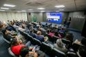 #ParaQuemNaoPodeVer Imagem de uma sala de conferências. Na foto, várias pessoas estão sentadas em poltronas, formando fileiras, e assistindo a uma apresentação feita por duas pessoas na frente da sala, próximo a um telão. No telão, há um slide azul com informações (textos e gráficos) e, na parede, o logotipo “Portos do Paraná” está visível. Também estão presentes bandeiras do Brasil.