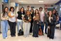#ParaQuemNaoPodeVer Grupo de onze jovens alinhados em um corredor interno, todos segurando crachás verdes voltados para a frente. Na foto, as pessoas estão em pé, lado a lado, sorrindo. Há diversidade de tons de pele, de claros a médios e mais escuros, e estilos de cabelo diferentes, incluindo cabelos lisos, cacheados e ondulados, alguns longos, outros curtos, em cores como castanho, preto e loiro. As roupas são casuais: calças jeans claras e escuras, calça de moletom cinza, calça preta, tênis brancos