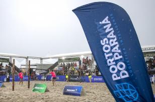 #ParaQuemNaoPodeVer A imagem mostra uma fotografia de um evento esportivo ao ar livre, possivelmente um torneio de vôlei de praia. No primeiro plano, há uma bandeira azul com o texto "PORTOS DO PARANÁ LOGÍSTICA INTELIGENTE". Ao fundo, é possível ver uma quadra de areia com jogadores vestindo uniformes rosa, além de várias pessoas assistindo sob uma estrutura coberta. Há também outras bandeiras e faixas com logotipos e textos, mas não estão totalmente legíveis na imagem.