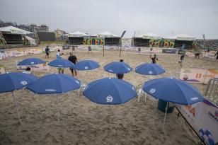 #ParaQuemNaoPodeVer A imagem mostra uma foto de uma praia. Na foto, há várias cadeiras de praia com guarda-sóis azuis, todos com o logotipo da "Portos do Paraná" estampado. Ao fundo, há algumas pessoas caminhando na areia e estruturas como tendas e banners, sugerindo que pode estar ocorrendo algum evento ou competição. Além disso, há prédios visíveis ao fundo, indicando que a praia está em uma área urbana.