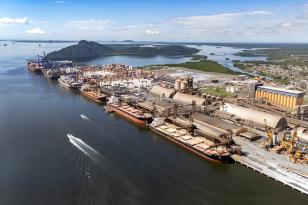 #ParaQuemNaoPodeVer Foto exibe uma imagem aérea de uma área portuária. A imagem mostra uma grande instalação industrial ao longo de um corpo d'água, com várias estruturas e edifícios. Há um barco deixando um rastro na água à esquerda. No fundo, é possível ver uma ponte e uma área montanhosa.