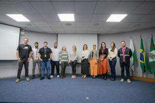 #ParaQuemNaoPodeVer Grupo de pessoas posando para uma foto em uma sala de reuniões. São 11 pessoas. Ao fundo, há três bandeiras: a do Brasil ao centro e duas verdes com detalhes vermelhos e um brasão, provavelmente de um estado ou município. O chão é acarpetado de azul e o teto tem placas brancas com iluminação embutida. As pessoas estão sorrindo e parecem estar em um evento formal ou institucional. 