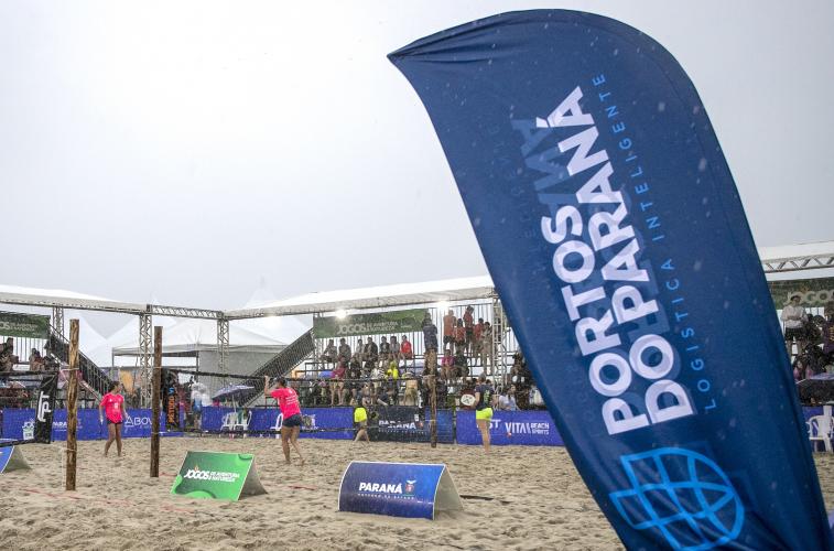 #ParaQuemNaoPodeVer A imagem mostra uma fotografia de um evento esportivo ao ar livre, possivelmente um torneio de vôlei de praia. No primeiro plano, há uma bandeira azul com o texto "PORTOS DO PARANÁ LOGÍSTICA INTELIGENTE". Ao fundo, é possível ver uma quadra de areia com jogadores vestindo uniformes rosa, além de várias pessoas assistindo sob uma estrutura coberta. Há também outras bandeiras e faixas com logotipos e textos, mas não estão totalmente legíveis na imagem.