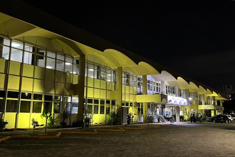 #ParaQuemNaoPodeVer Imagem exibe uma fotografia de um prédio iluminado à noite. O prédio tem uma fachada com muitas janelas e está iluminado com luzes amarelas, criando um efeito visual marcante. A imagem está inclinada, mostrando a lateral do prédio, que parece ter uma arquitetura moderna com formas curvas.