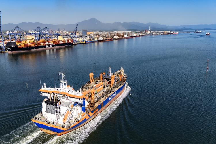 Uma draga navega pelo canal marítimo de acesso ao Porto de Paranaguá. A embarcação, nas cores branca, laranja e azul, aparece em primeiro plano deixando um rastro de espuma na água, indicando movimentação. Ao fundo, é possível ver a estrutura do Porto de Paranaguá, com guindastes alinhados, contêineres empilhados e um navio cargueiro atracado. Mais atrás, surgem as montanhas da Serra do Mar sob uma iluminação suave, característica do início da manhã ou do fim da tarde.