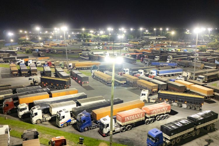 Imagem aérea noturna Pátio de Triagem de caminhões da Portos do Paraná. Vários caminhões e carretas estão estacionados lado a lado sob uma intensa luz que sai dos postes e super postes.