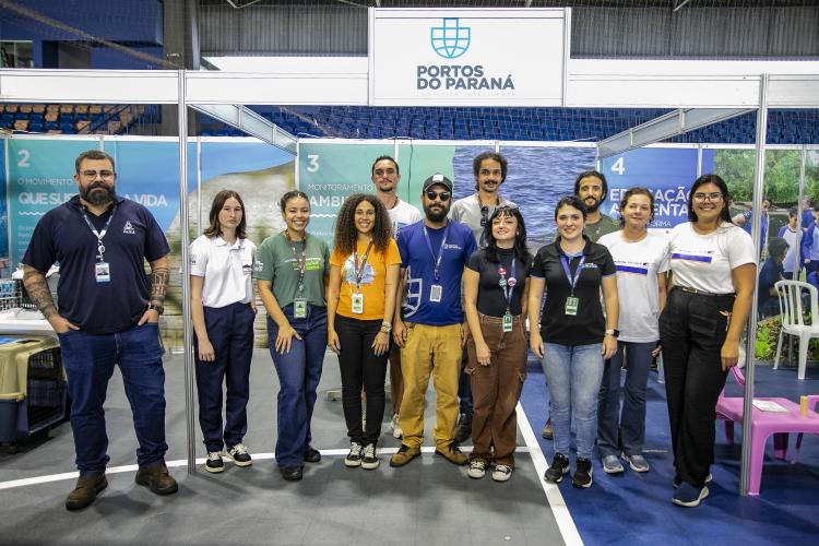 Há onze pessoas em pé, alinhadas em três fileiras, todas sorrindo para a câmera. Todos fazem parte da equipe de Meio Ambiente da Portos do Paraná. Eles estão posicionados na frente do stand da empresa.