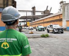 #ParaQuemNaoPodeVer A imagem mostra um homem em primeiro plano, visto de costas, usando um capacete de segurança cinza e uma camisa verde. Ao fundo, há uma estrutura industrial com um prédio laranja e azul e duas viaturas. A primeira, traz na carroceria a imagem de Nossa Senhora do Rocio. Atrás das viaturas, seguem outros veículos em fila, acompanham o cortejo pelas instalações do Porto de Paranaguá.