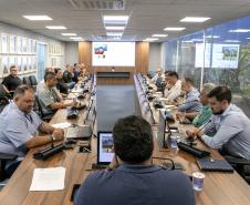 #ParaQuemNaoPodever Na foto, há várias pessoas sentadas ao redor de uma longa mesa de conferência. As pessoas estão usando roupas formais e parecem estar participando de uma reunião ou discussão. Há microfones na mesa em frente a cada pessoa, e algumas delas estão usando laptops ou tablets. A sala tem uma parede de vidro ao fundo, através da qual é possível ver plantas. Na parede oposta, há quadros com retratos de pessoas. A iluminação é feita por lâmpadas embutidas no teto.