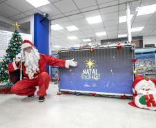 #ParaQuemNaoPodeVer A imagem mostra um homem vestido de Papai Noel agachado ao lado de uma árvore de Natal decorada com bolas azuis e douradas e uma estrela dourada no topo. Ele está usando um traje vermelho com detalhes brancos, incluindo um gorro e uma longa barba branca. O homem está fazendo um gesto de "paz e amor" com a mão direita. Ao lado dele, há um painel azul com a inscrição "Natal Solidário" em letras brancas e uma estrela amarela acima do texto. Na frente do painel, há um tapete ou almofada com 