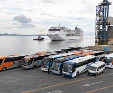 #praquemnaopodever 7 ônibus e uma van estão a frente da foto, no cais do Porto de Paranaguá, aguardando a chegada, atrás, do imponente navio de cruzeiros MSC Armonia. 