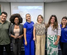 #ParaQuemNaoPodeVer A imagem mostra seis pessoas posando para uma foto em um ambiente interno. Há um homem de camisa verde e duas mulheres, uma com cabelo afro e outra com cabelo preso. Na fileira da frente, há três mulheres, uma usando uma camisa azul, outra com uma blusa estampada e a terceira com uma blusa branca e saia com listras verdes. Ao fundo, há bandeiras e um painel que parece ter um mapa ou uma imagem de cidade.