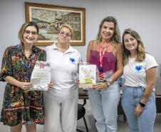 #ParaQuemNaoPodeVer A imagem mostra quatro pessoas posando para uma foto em um ambiente interno. Duas mulheres estão em pé na parte de trás, uma delas usando um vestido estampado colorido e óculos, segurando um papel, e a outra usando uma blusa branca. Na frente, há duas mulheres, uma usando uma blusa rosa e segurando um pacote, e a outra com uma camiseta branca com um desenho azul. Ao fundo, há uma pintura emoldurada na parede.