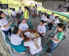 #praquemnaopodever várias pessoas sentadas em cadeiras de plástico verdes separadas em grupos gesticulam sobre projetos socioambientais.  Todos vestem camisas brancas e conversam sérios enquanto outras pessoas em pe passam pelas pesas. Um prancha está presa ao teto e ao fundo é possível vera grama da Ilha do Teixeira